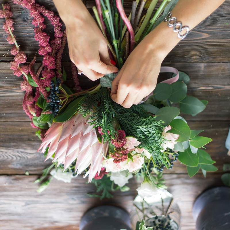 Bouquet Protée : composition florale rustique de mariage. Mains de fleuriste attachant un bouquet rustique avec une grande Protée rose, Amarante rouge, Eucalyptus et baies sur une table en bois.