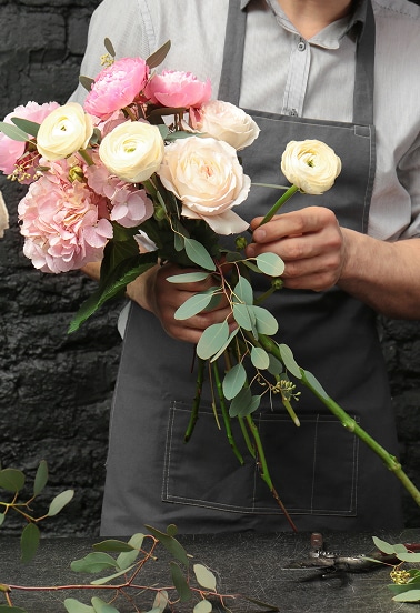 Bouquet de roses et renoncules: l'art du fleuriste Mains d'un fleuriste composant un bouquet de roses, renoncules crème et hortensias roses avec de l'eucalyptus frais sur fond sombre.
