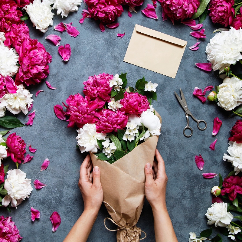 Pivoines roses et blanches : Créer un bouquet raffiné Bouquet vibrant de pivoines fuchsia et blanches, enveloppé dans du kraft. Pétales, ciseaux et enveloppe sur fond gris foncé.