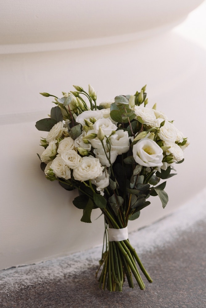 Bouquet de Mariée Blanc : Roses, Lisianthus et Eucalyptus Bouquet de roses blanches, lisianthus et eucalyptus élégant avec ruban blanc contre un mur texturé.